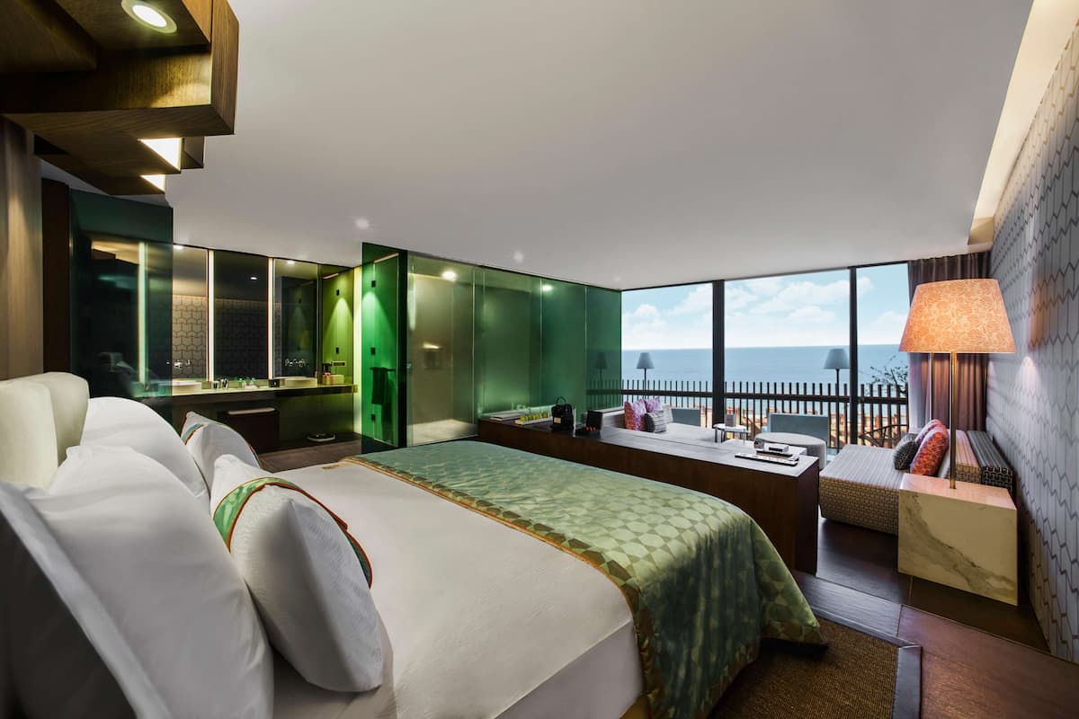 A modern hotel room with a large bed, a desk, and a view of the sea.