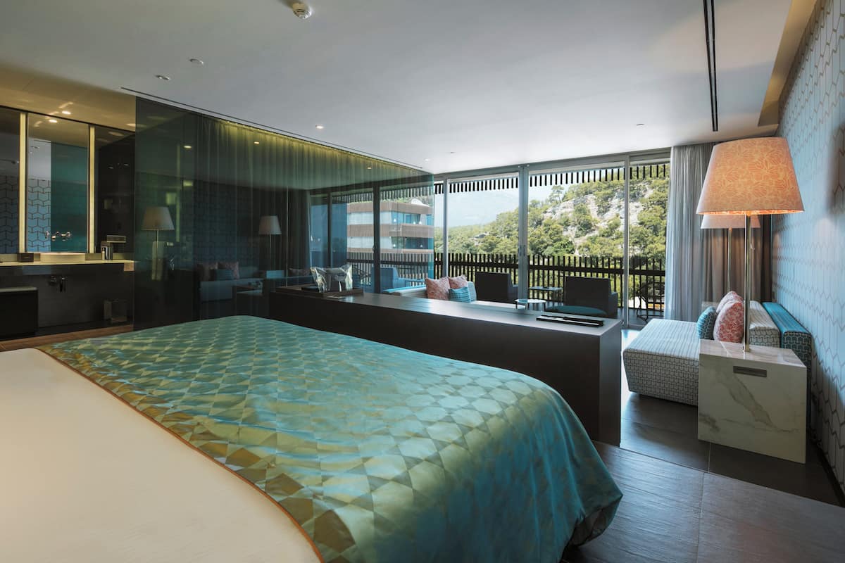 A modern hotel room with a large bed, a seating area with a view, a bathroom with a glass partition, and a balcony with a view.