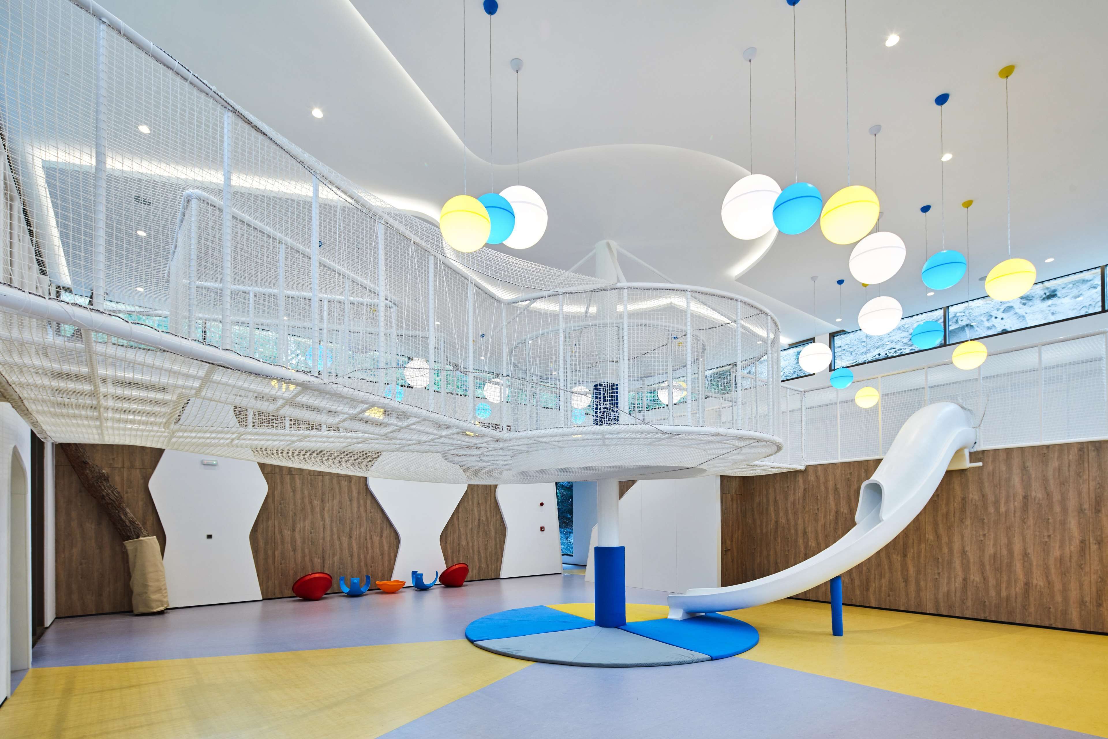 A modern indoor play area with a slide, colorful hanging lights, and a netted play structure.