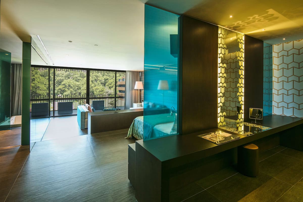 A modern hotel room with a large window, a bed, a desk, and a seating area.