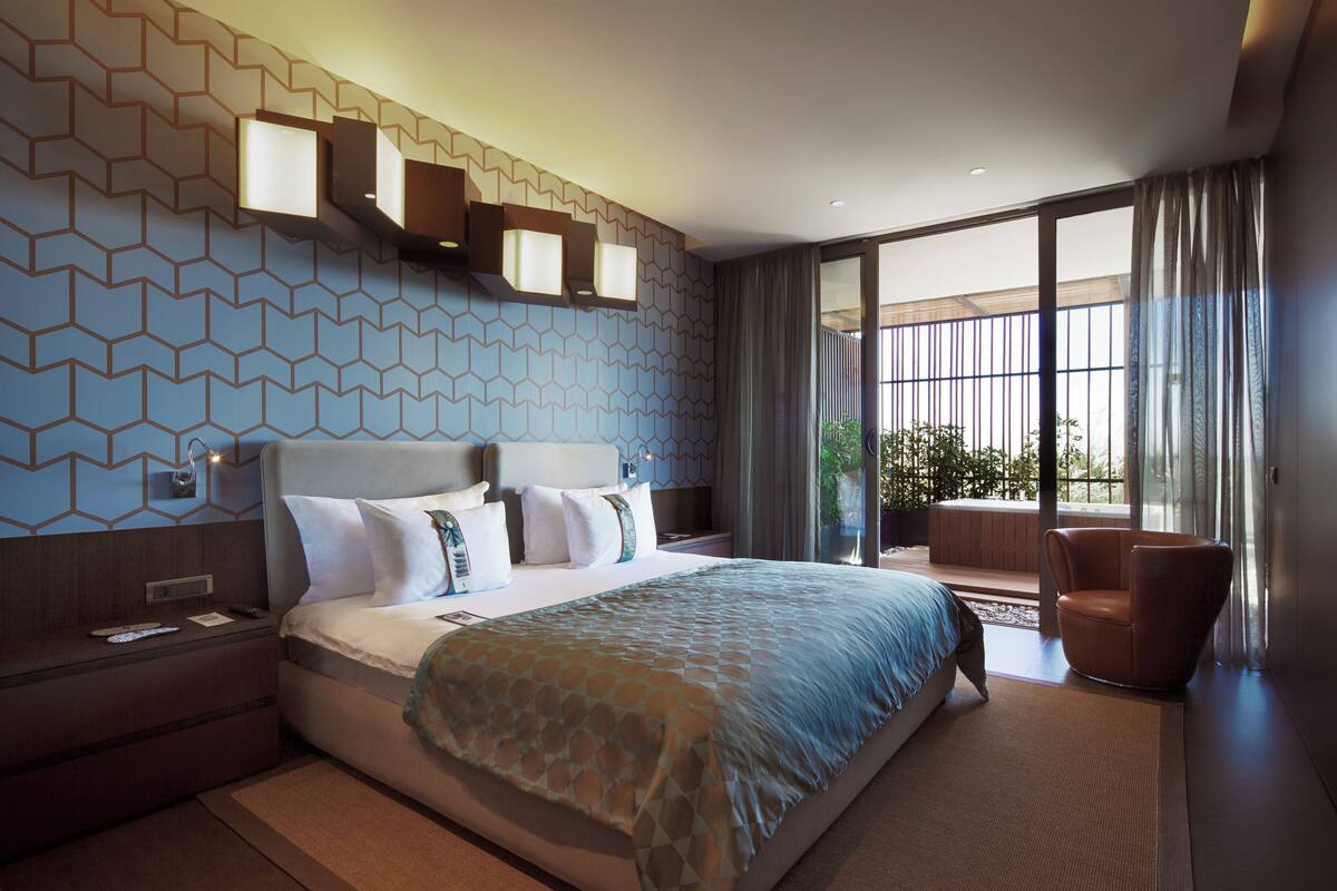 A modern hotel room with a large bed, a seating area with a chair, and a view of the outdoors through large windows.