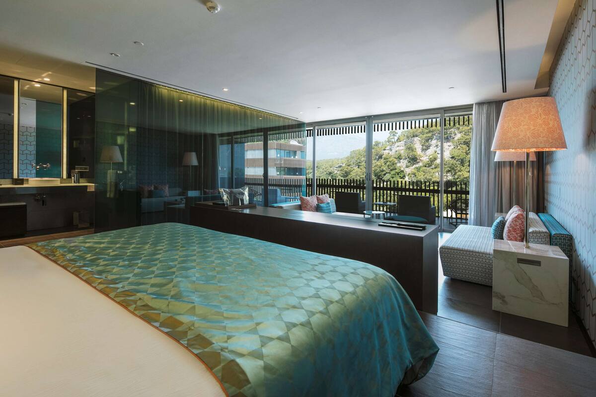 A modern hotel room with a large bed, a seating area with a view, a bathroom with a glass partition, and a balcony with a railing.