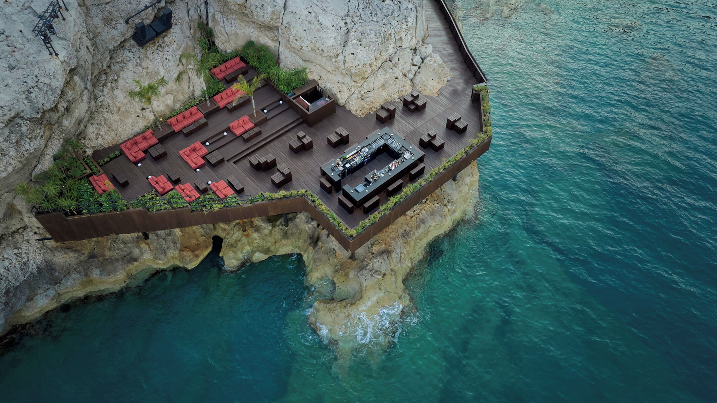 Luchtbeeld van een resort aan de klif met een houten terras, rode ligbedden en een bar.
