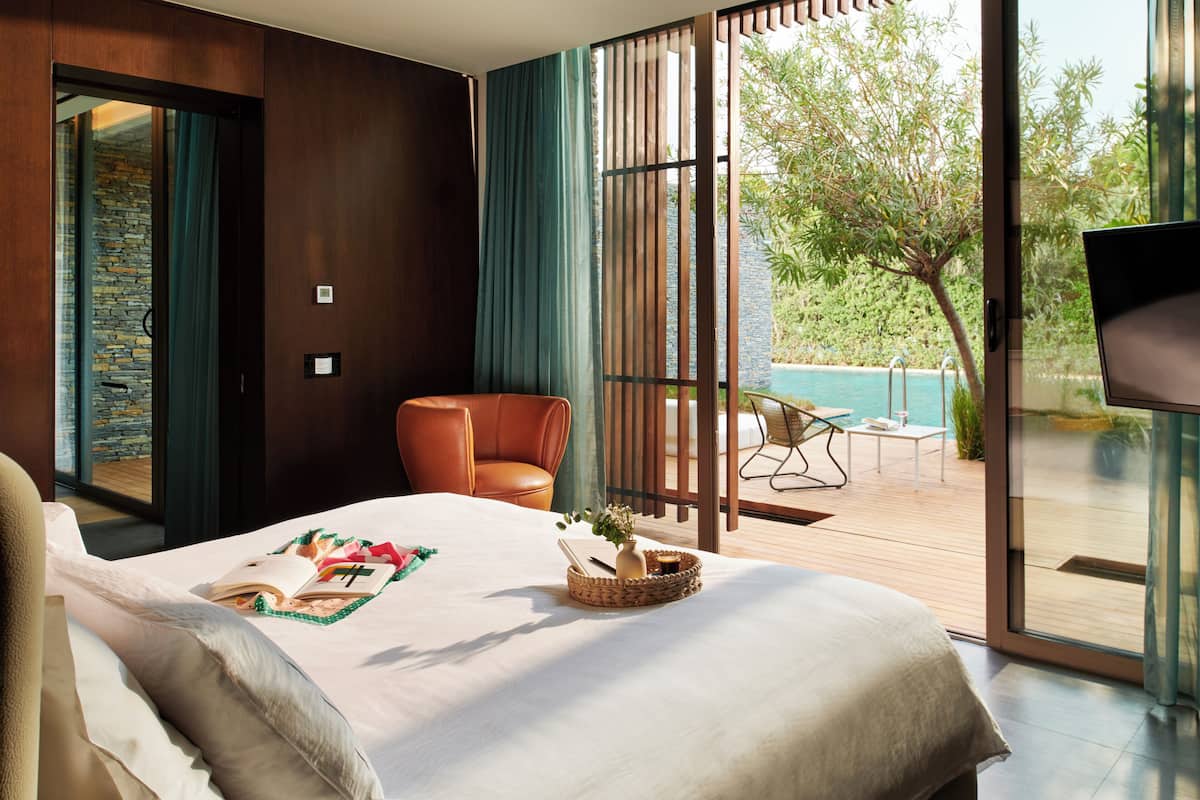 A modern hotel room with a large bed, a seating area with a chair and table, and a view of an outdoor pool and greenery.