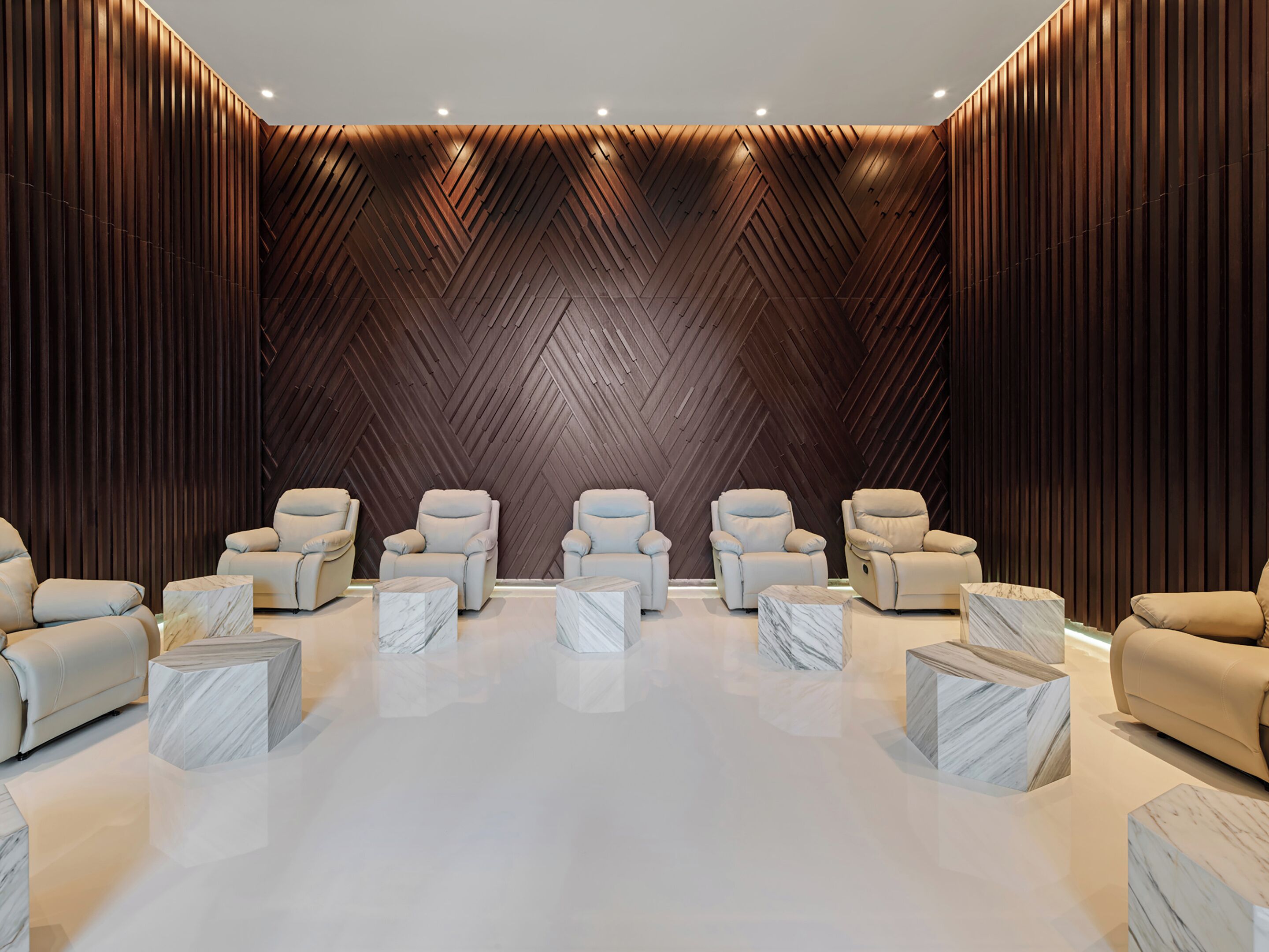 Un spa moderno con seis sillas de masaje beige dispuestas en dos filas, suelo de mármol y pared de madera oscura con diseño en zigzag.