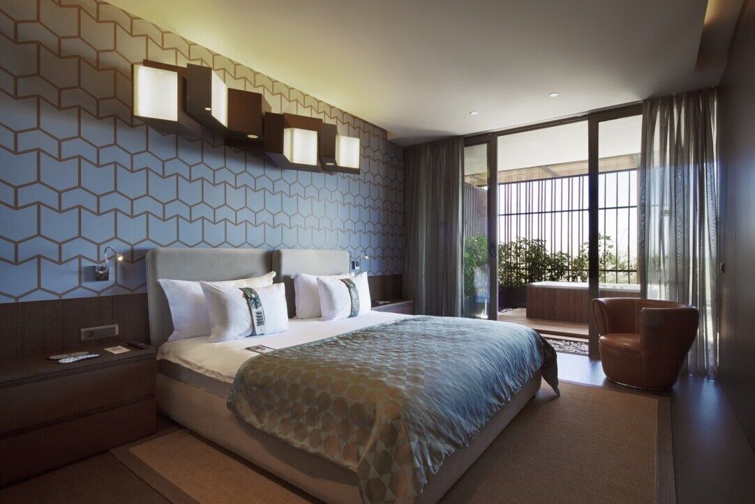 Habitación de hotel moderna con una cama grande, una mesita de noche, una zona de estar con una silla y vistas a zonas verdes a través de amplios ventanales.