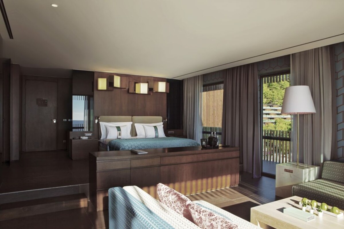 A modern hotel room with a large bed, a sofa, a coffee table, and a view of the outdoors.