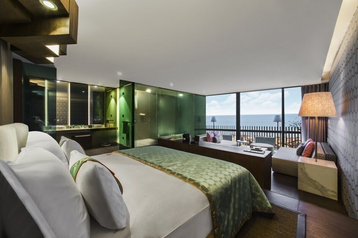A modern hotel room with a large bed, a seating area with a view, a vanity with a mirror, and a balcony with a railing.