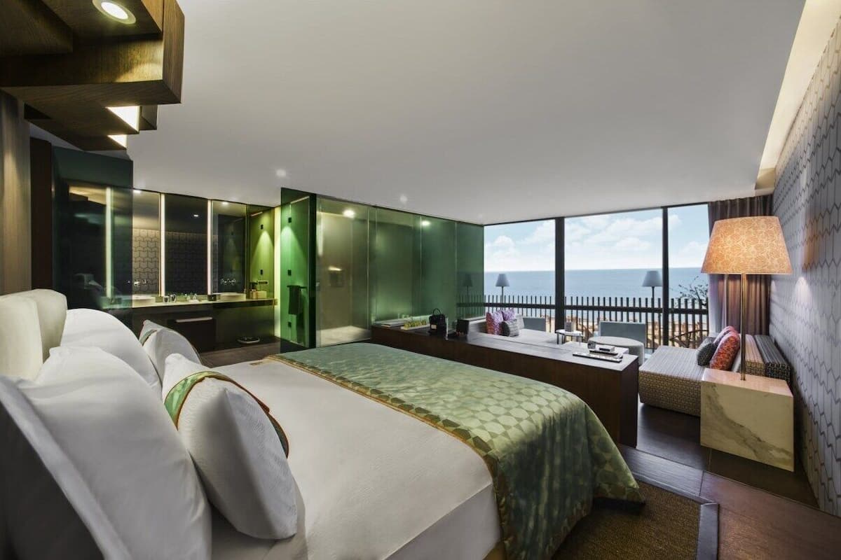 A modern hotel room with a large bed, a seating area with a view, a vanity with a mirror, and a balcony with a railing.