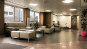Un vestíbulo amplio con un sofá grande, sillones y una mesa de centro. Se observan plantas en macetas y una estantería al fondo.