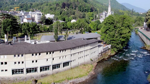 Un edificio a orillas del río, de estilo arquitectónico moderno, rodeado de abundante vegetación y montañas al fondo.