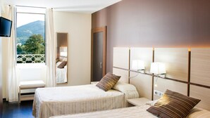 Habitación de hotel con dos camas, un balcón y vistas a la montaña, un televisor y una mesita de noche con lámparas.