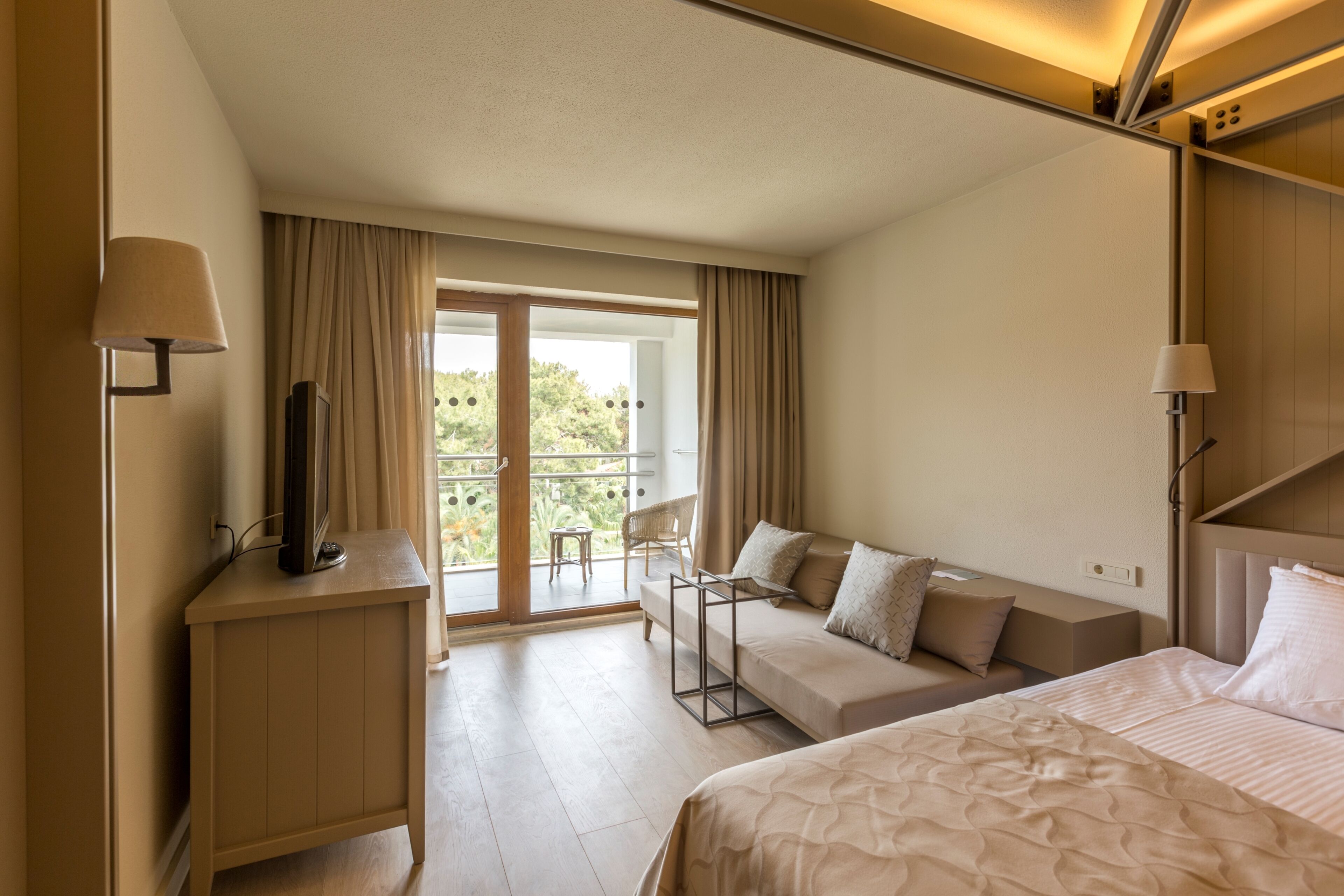 A hotel room with a bed, a sofa, a TV on a stand, a desk, and a balcony with curtains.