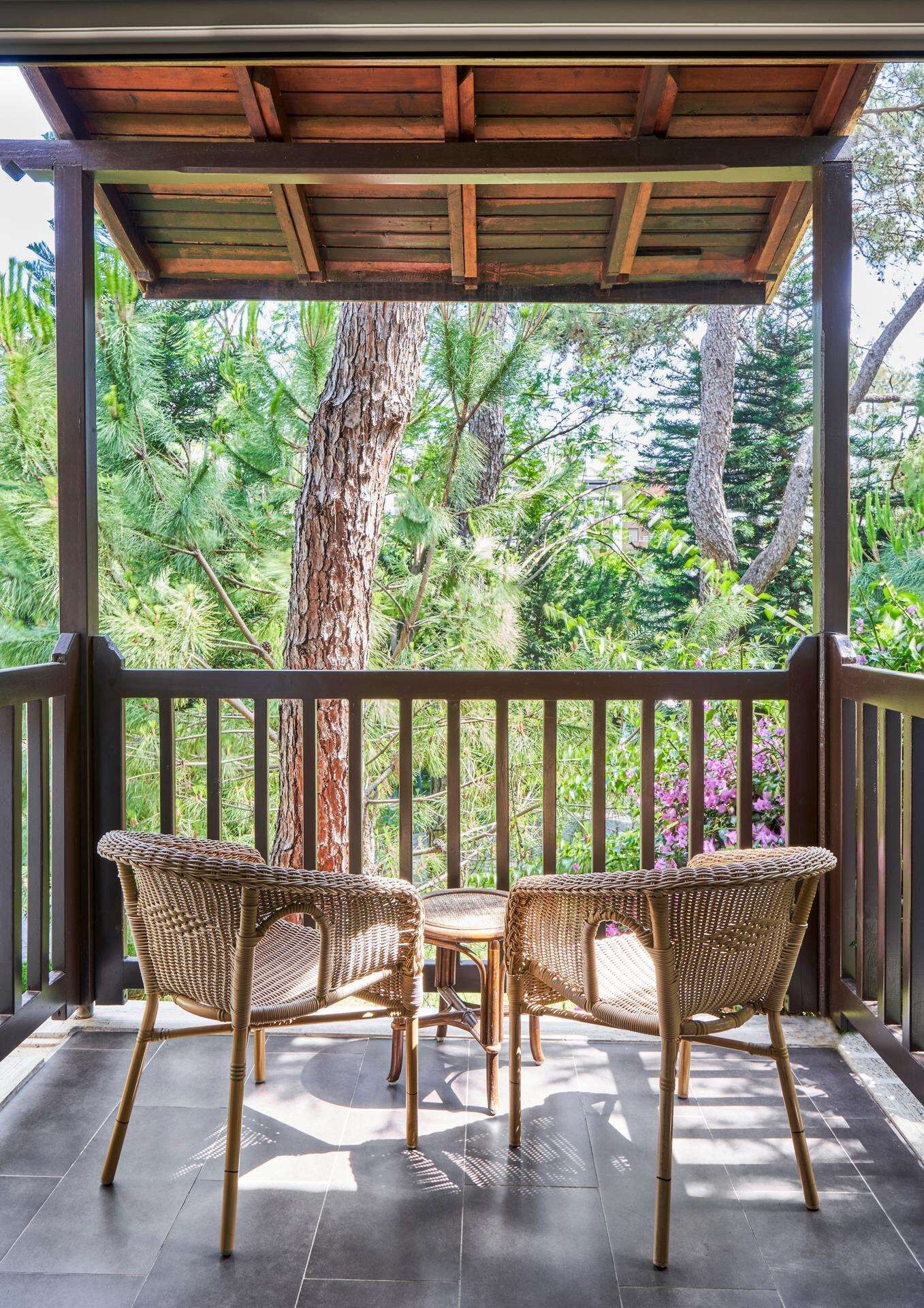Un espacio de estar al aire libre cubierto con dos sillas de mimbre y una mesa, rodeado por una barandilla de madera, con vistas a un jardín exuberante con árboles y plantas.