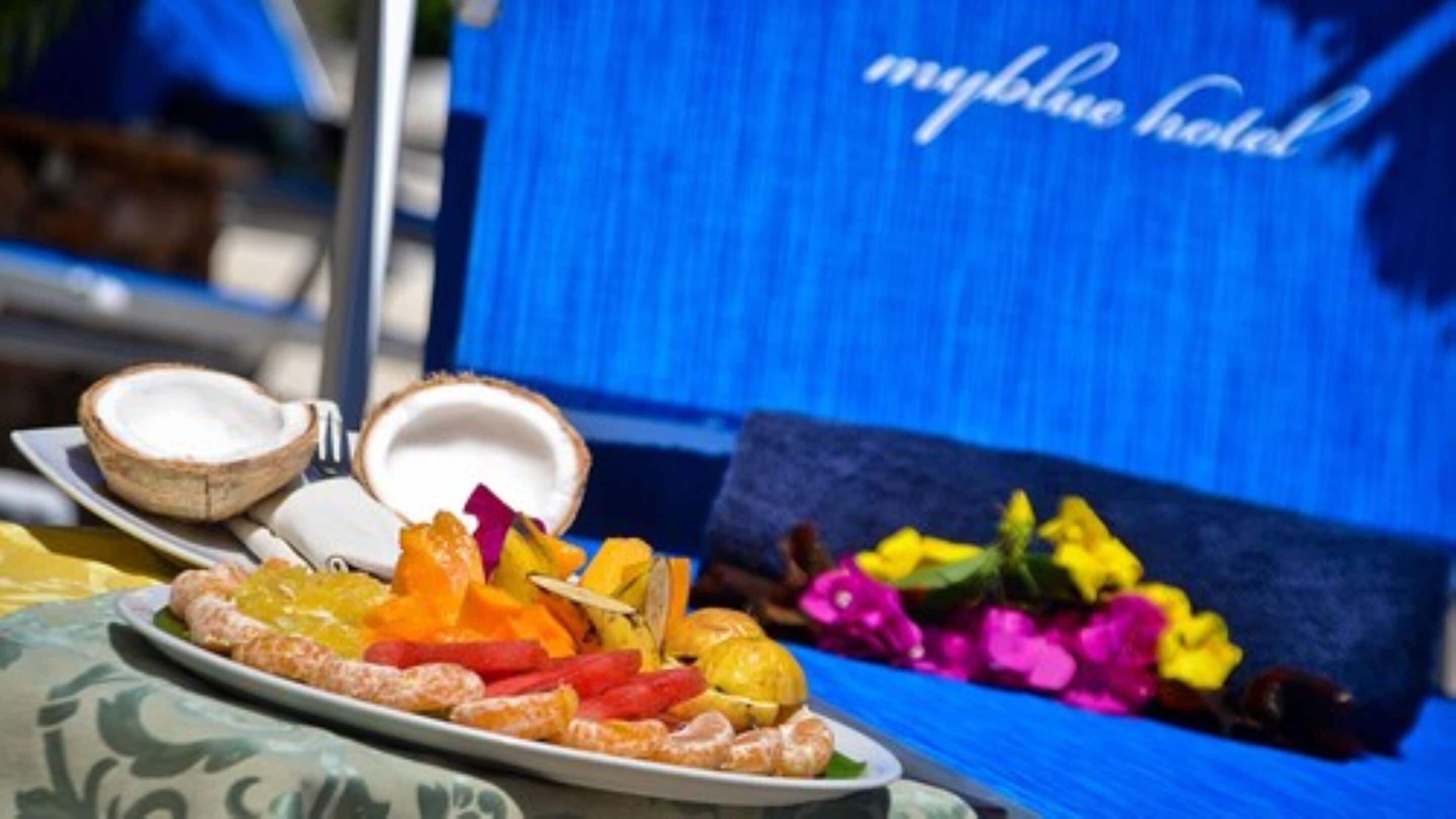 Une table garnie d’une variété de fruits et une assiette contenant un plat, le tout placé devant une chaise bleue sur laquelle on peut lire le texte « myblue hotel ».