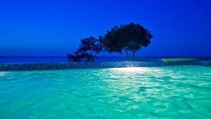 Una piscina con agua azul cristalina, rodeada por un borde de azulejos y un árbol al fondo.