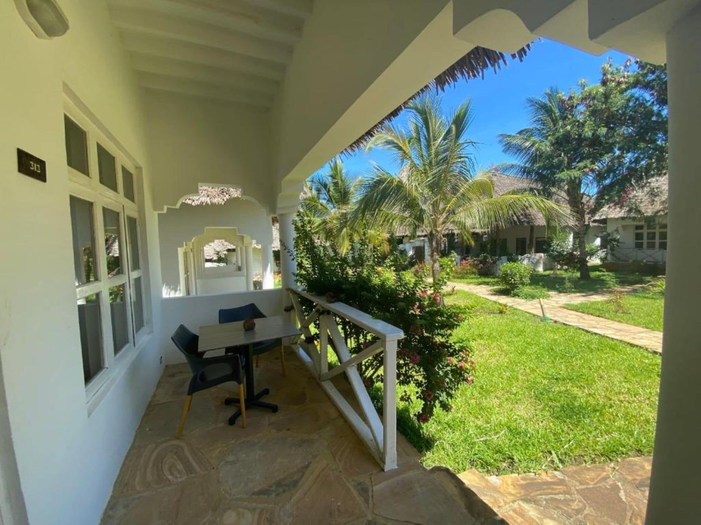 A covered outdoor area with a table and chairs, a stone floor, and a view of a garden with palm trees and a pathway.