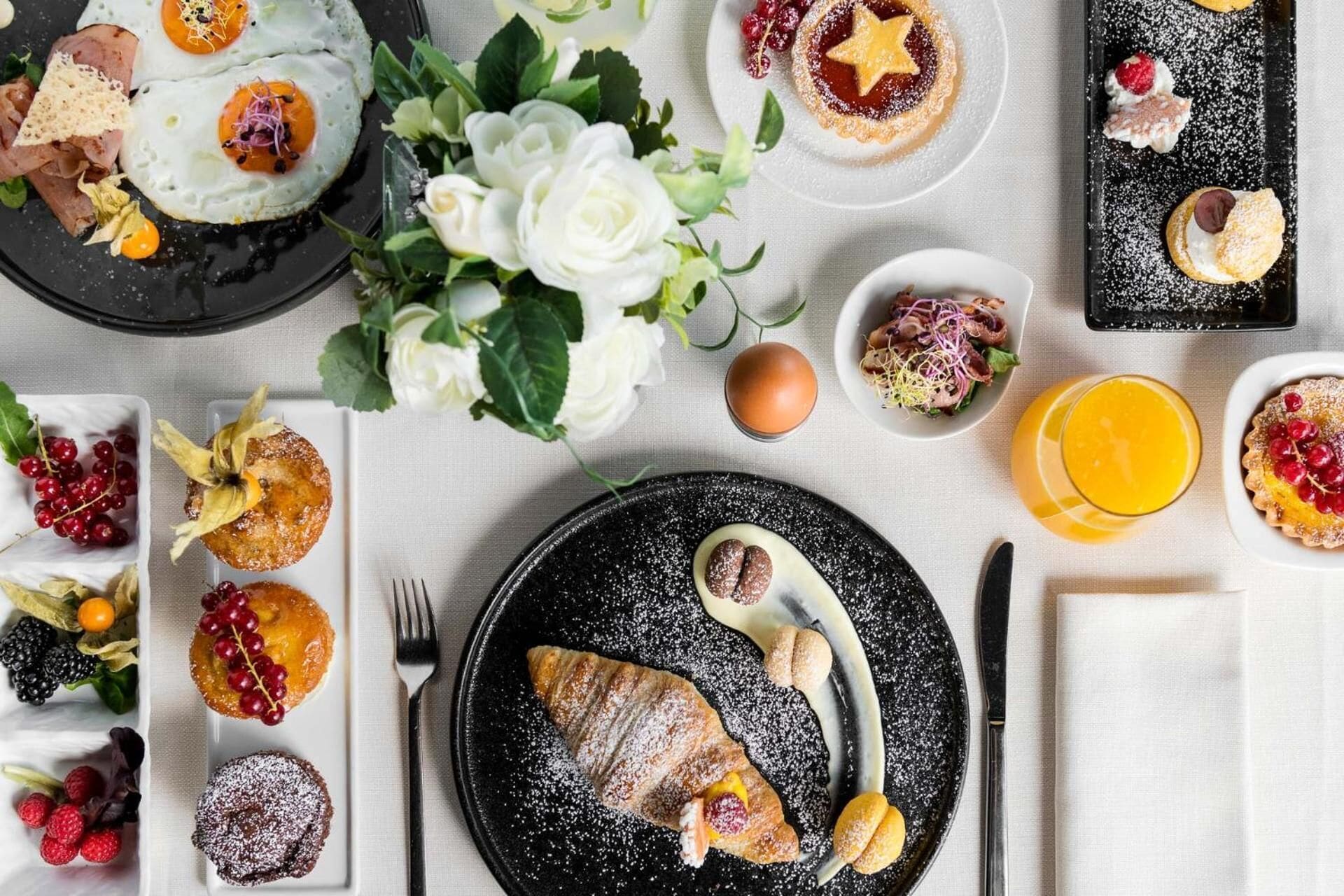 Una mesa dispuesta con diferentes platos, incluyendo huevos, pasteles y fruta, sobre un mantel con un jarrón de flores.