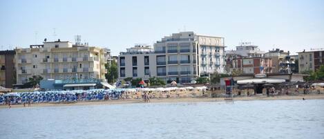Un paisaje urbano costero con una zona de playa que muestra hileras de sombrillas azules y tumbonas.
