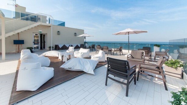 Terraza en la azotea con sillas de descanso blancas, una mesa de madera y sombrillas, con vista a un mar azul y despejado.