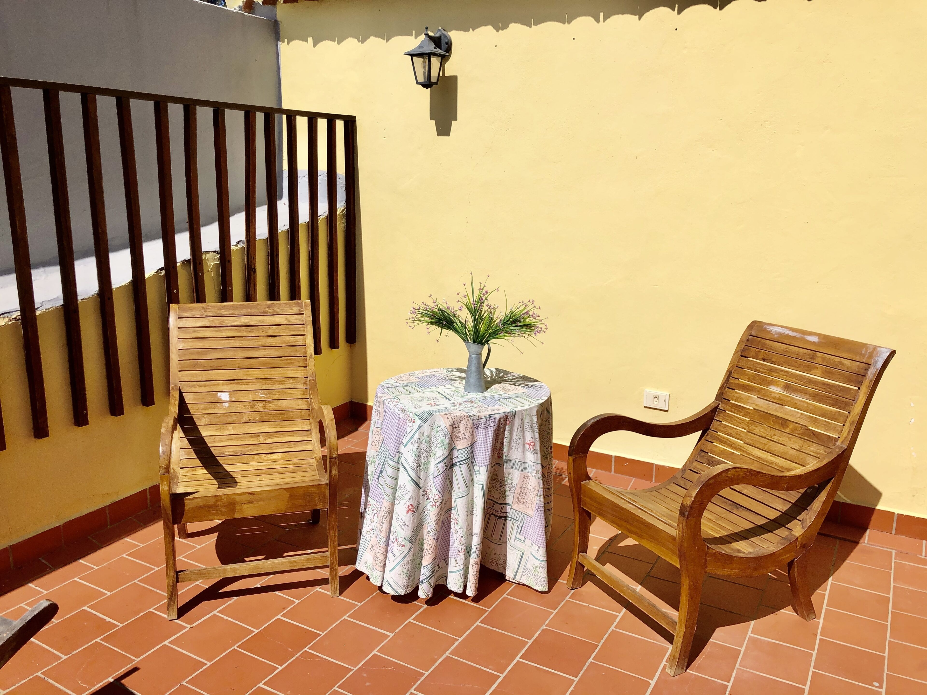 Een terras met twee houten stoelen, een kleine ronde tafel, een bloempot en een wandlamp.