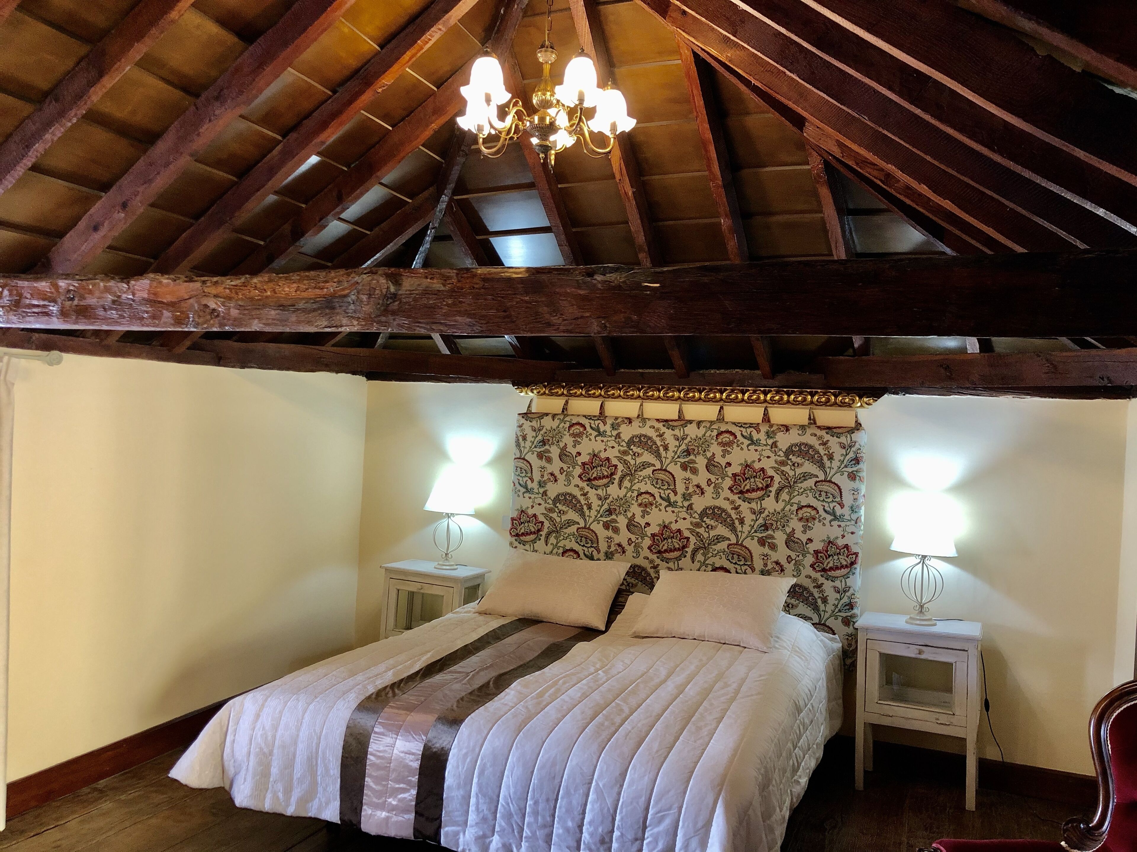 Une chambre avec un plafond en bois, un lit recouvert d’une couvre-lit à rayures, deux tables de chevet avec des lampes et une tête de lit à motifs floraux.