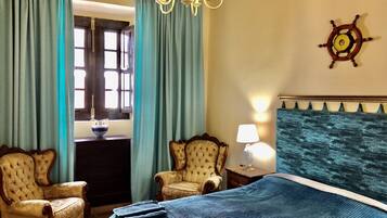 A hotel room with a bed, two armchairs, a chandelier, a ship's wheel decoration, and a window with curtains.