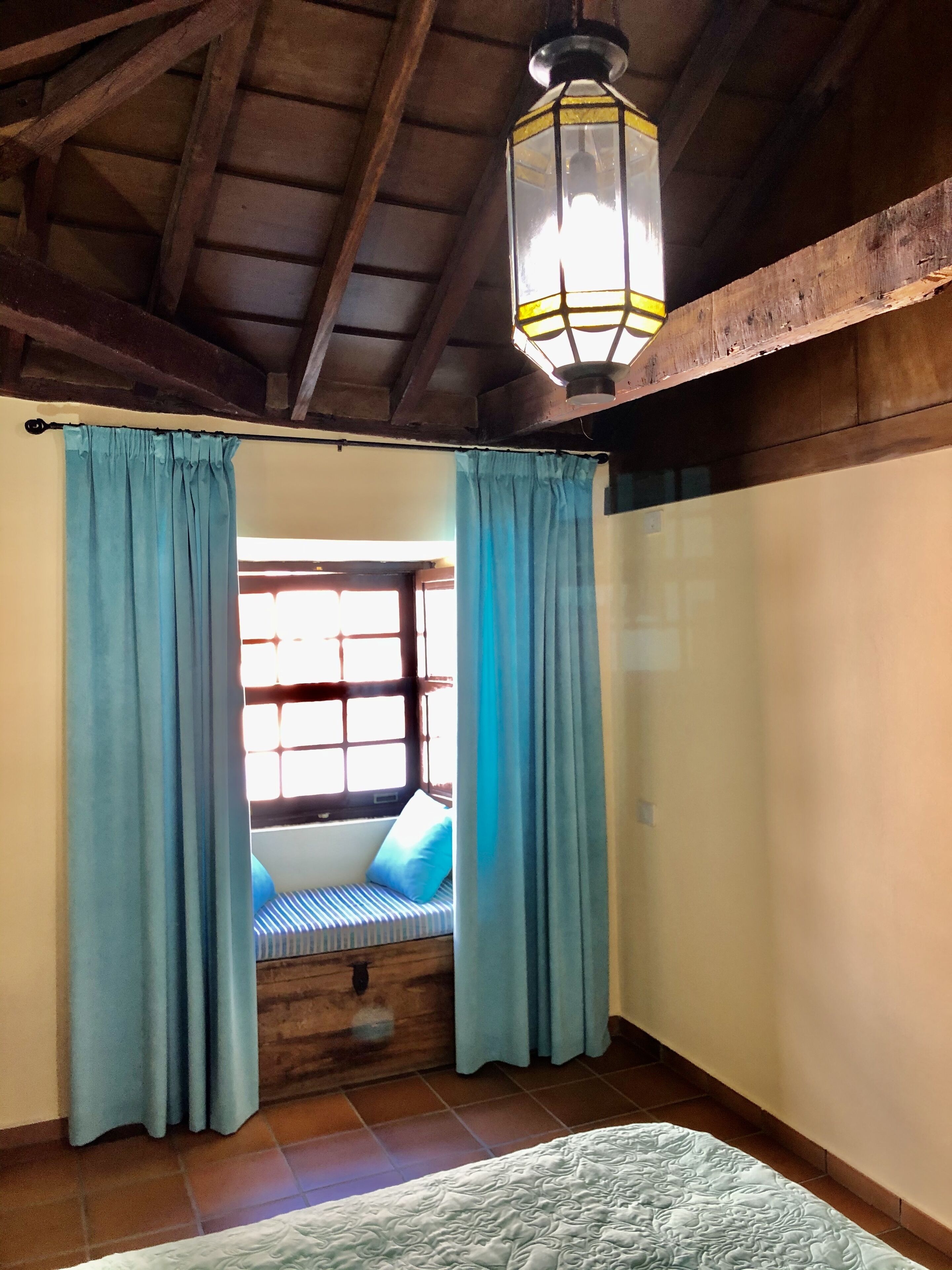 A room with a bed, a window with curtains, a hanging light fixture, and wooden beams on the ceiling.