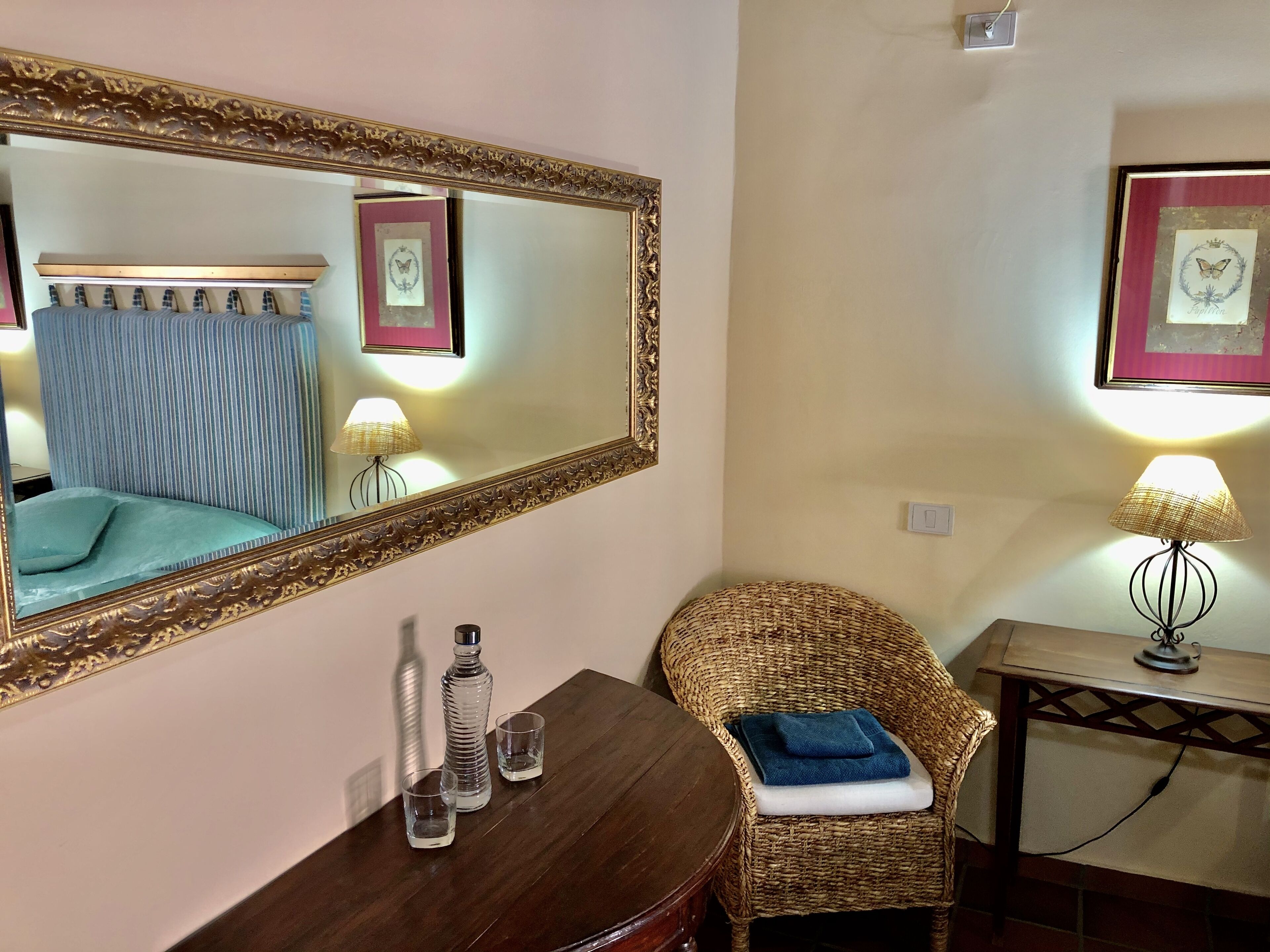Una habitación con un espejo grande, una silla de mimbre con una toalla, una mesita con una lámpara y una cama con cortinas a rayas.