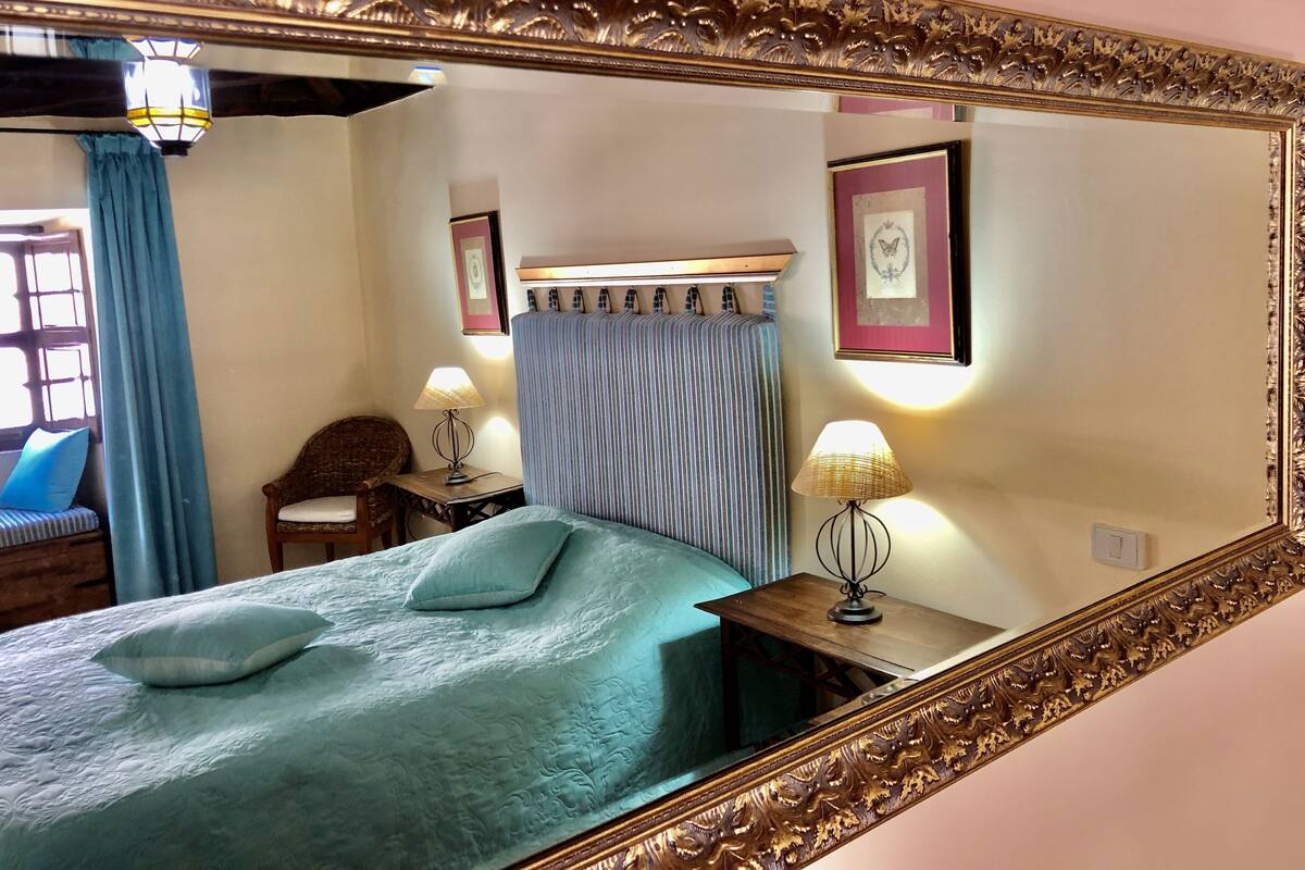 A bedroom with a large ornate mirror, a bed with a blue bedspread, a bedside table with a lamp, a chair, and a window with curtains.