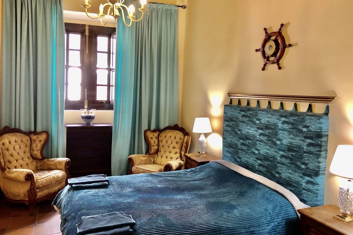 A bedroom with a large bed, two armchairs, a chandelier, a ship's wheel decoration, and a view of the outside through a window.
