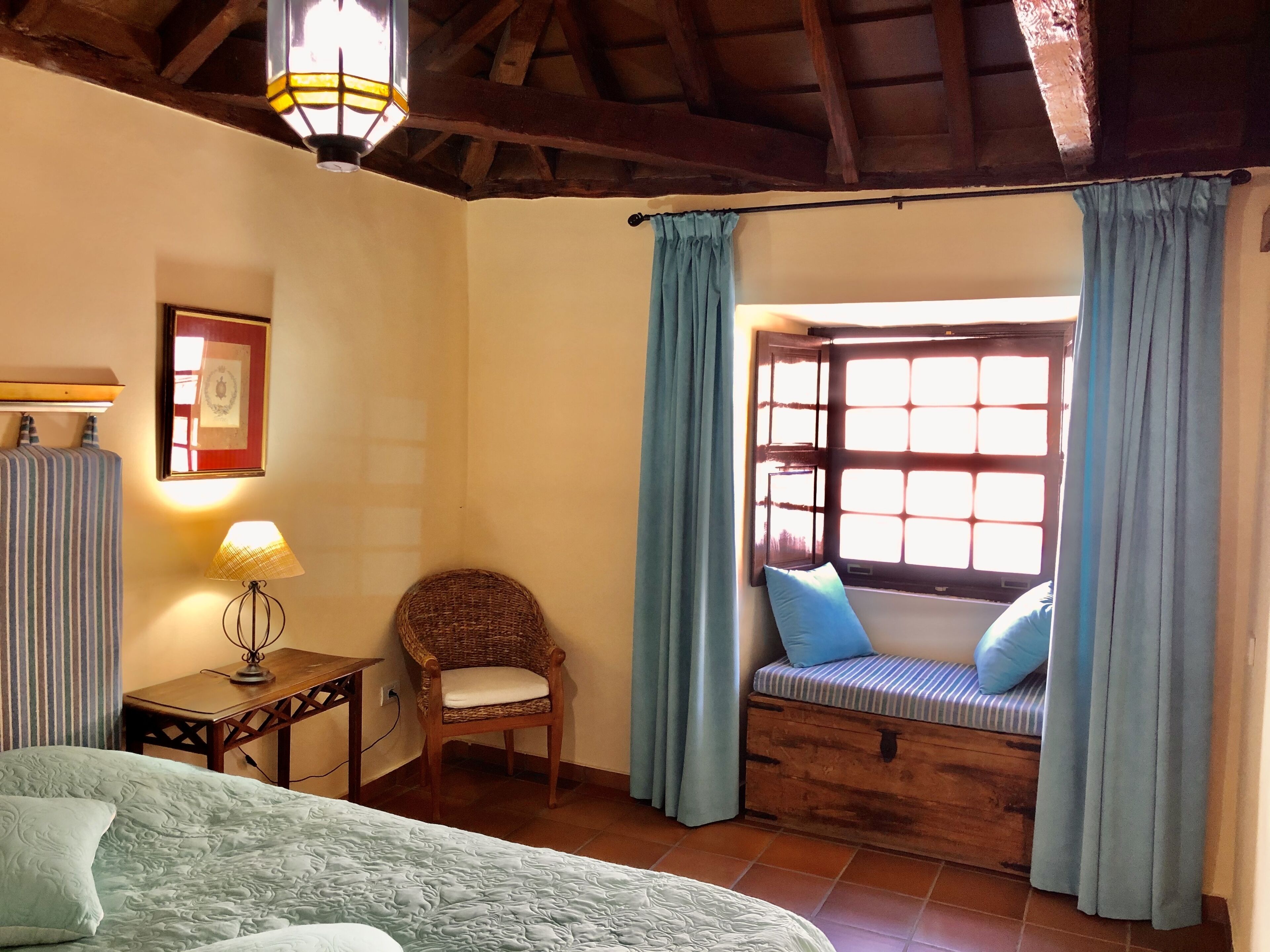 A cozy room with a bed, a wicker chair, a small table with a lamp, a window with blue curtains, and a framed picture on the wall.