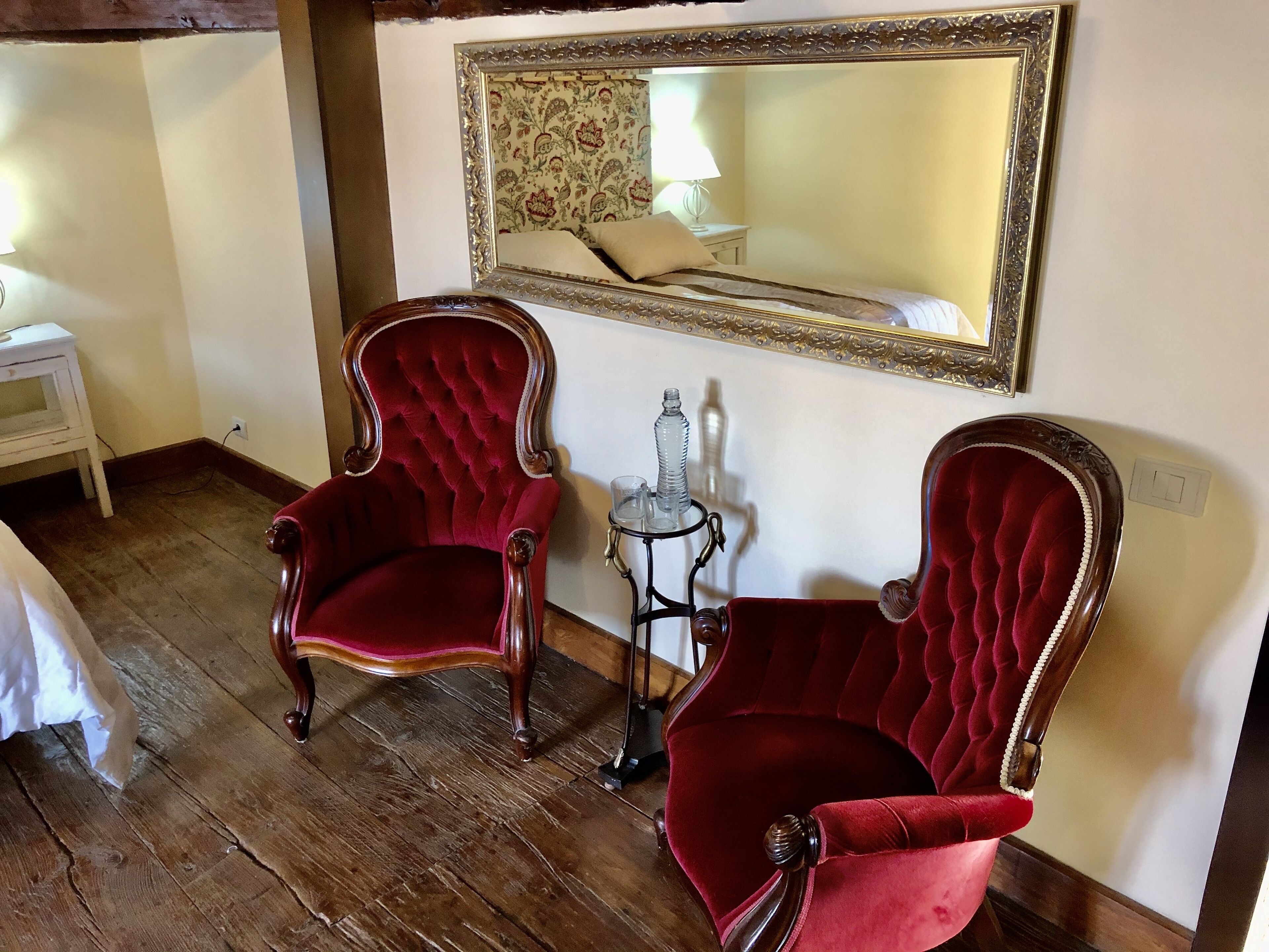 Um quarto com duas poltronas de veludo vermelho, uma pequena mesa com abajur e um espelho na parede.