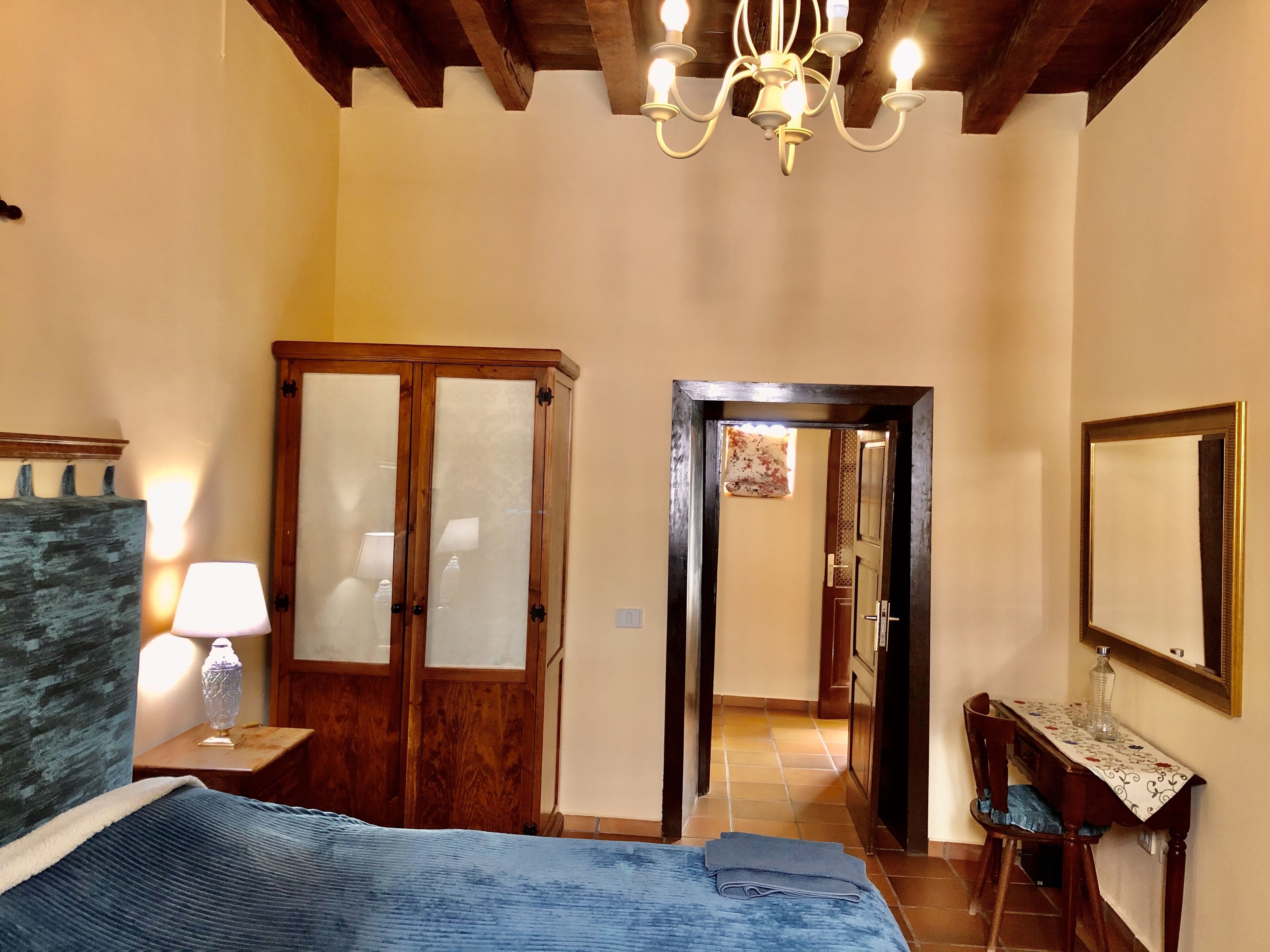A bedroom with a bed, a wooden wardrobe, a desk with a mirror, and a chandelier.