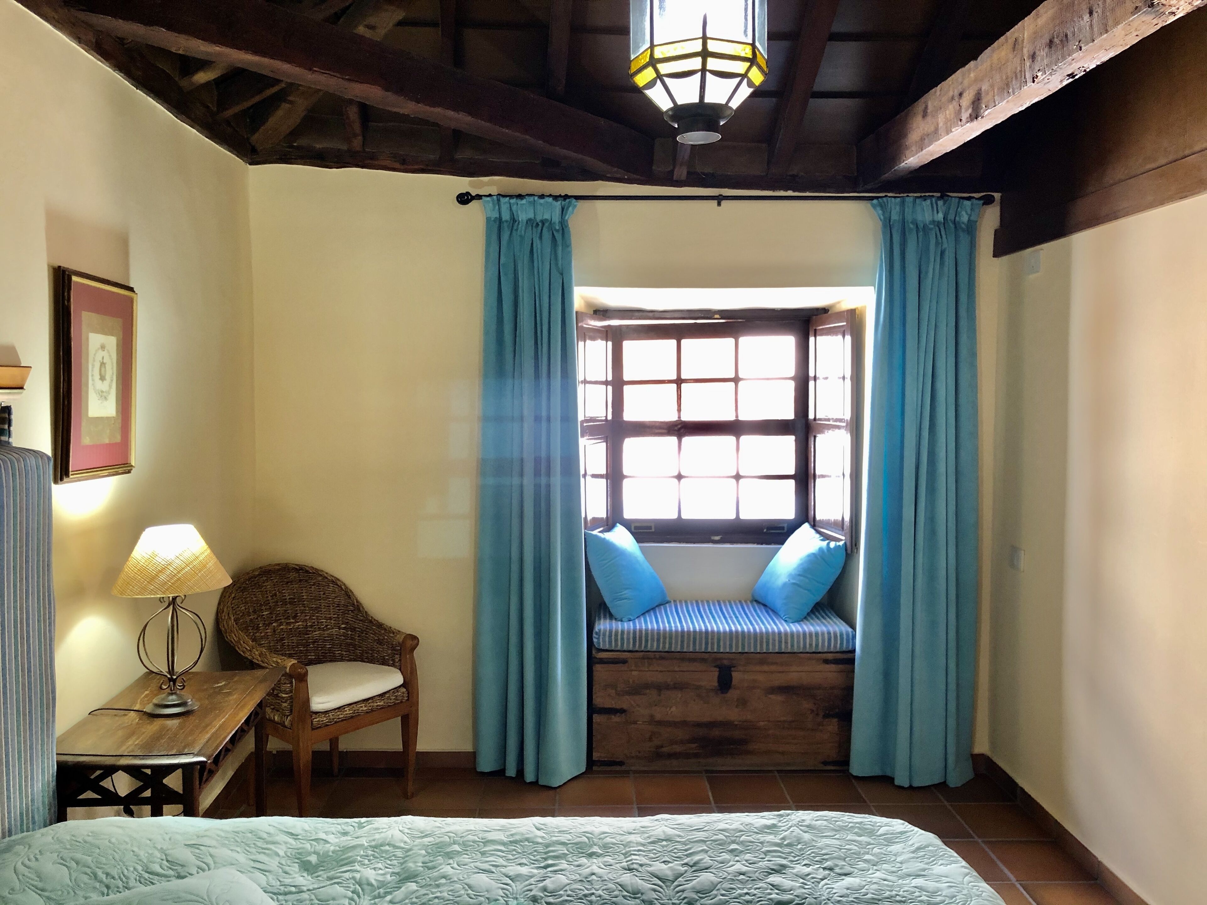 Una habitación con una cama, una silla de mimbre, una mesa con una lámpara, una ventana con cortinas azules y un banco de madera con cojines.