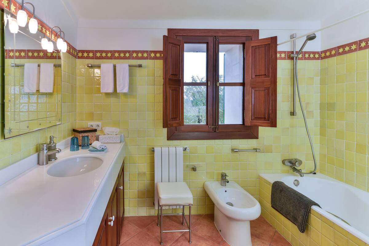 A bathroom with yellow tiles, a sink, a bidet, a bathtub, and a window with wooden shutters.