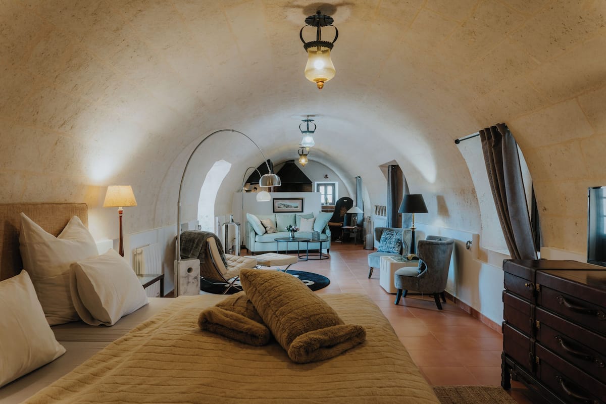 A cozy, well-lit room with a bed, a sofa, and a dresser. There is a lamp on the dresser and another on the nightstand. The room has a rustic, arched ceiling.