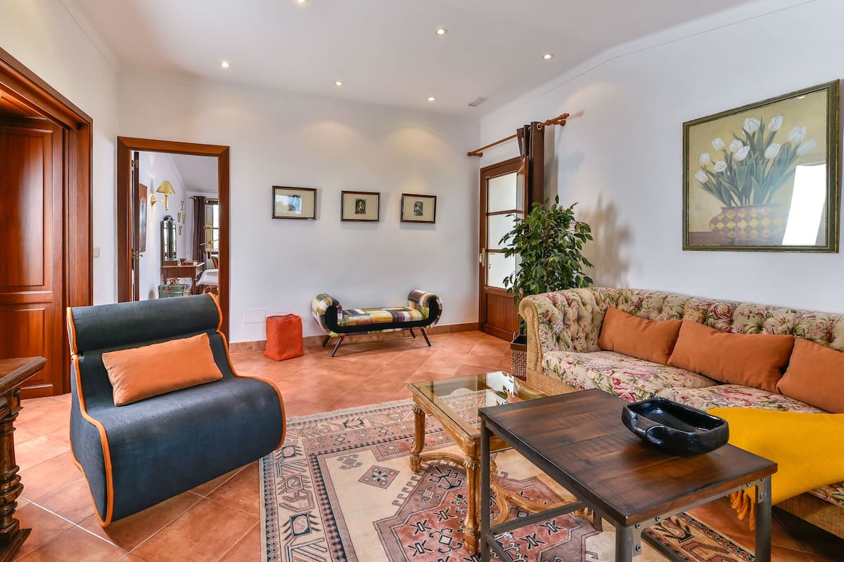 A living room with a blue armchair, a floral sofa, a glass coffee table, and a patterned rug.