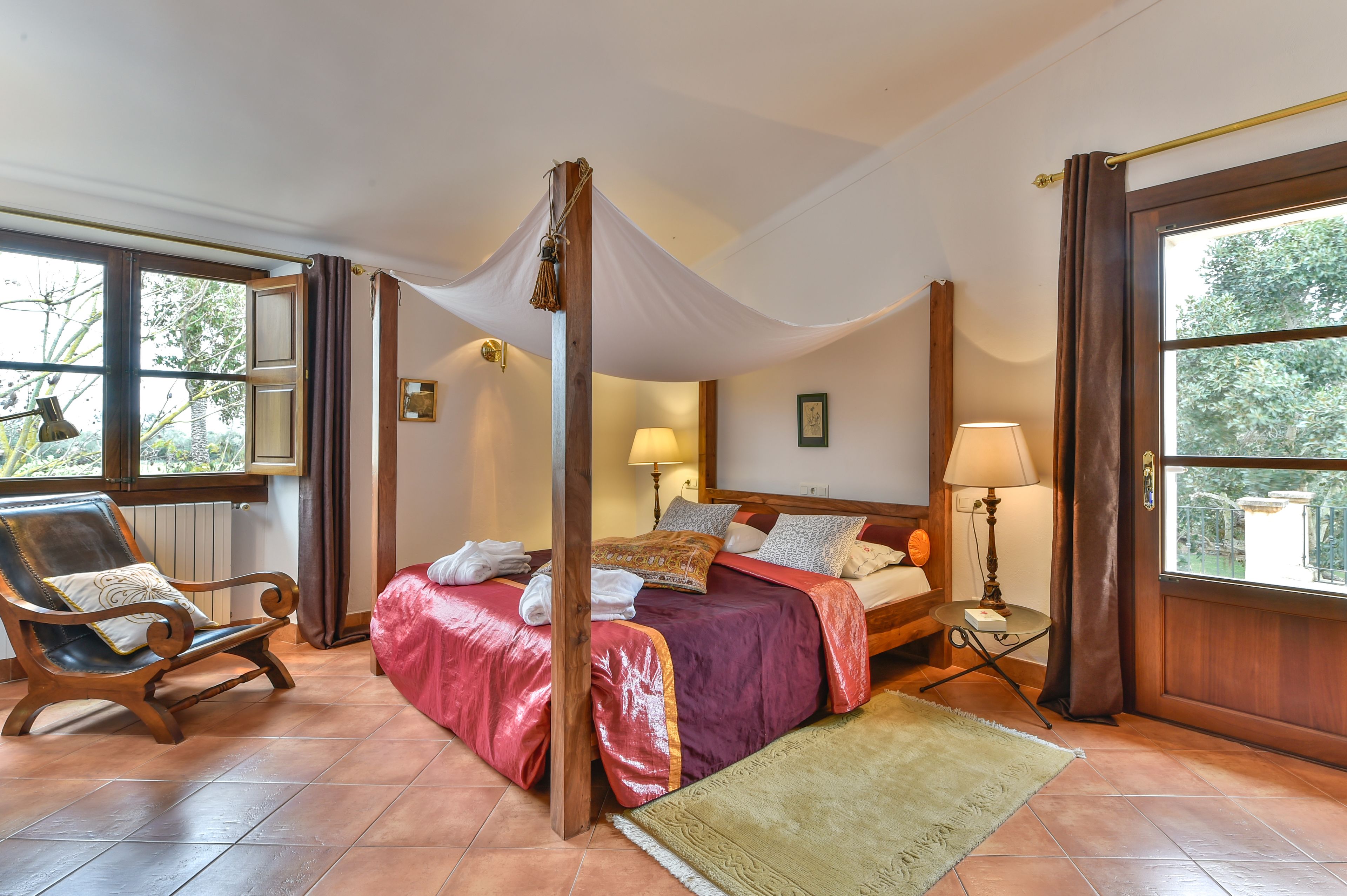 A four-poster bed with a canopy, a wooden chair with a cushion, a bedside table with a lamp, and a view of greenery through the windows.