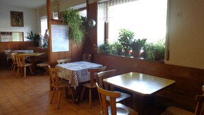 A restaurant with wooden tables and chairs, potted plants, and a window with curtains. - Garnì Astor (Alleghe)