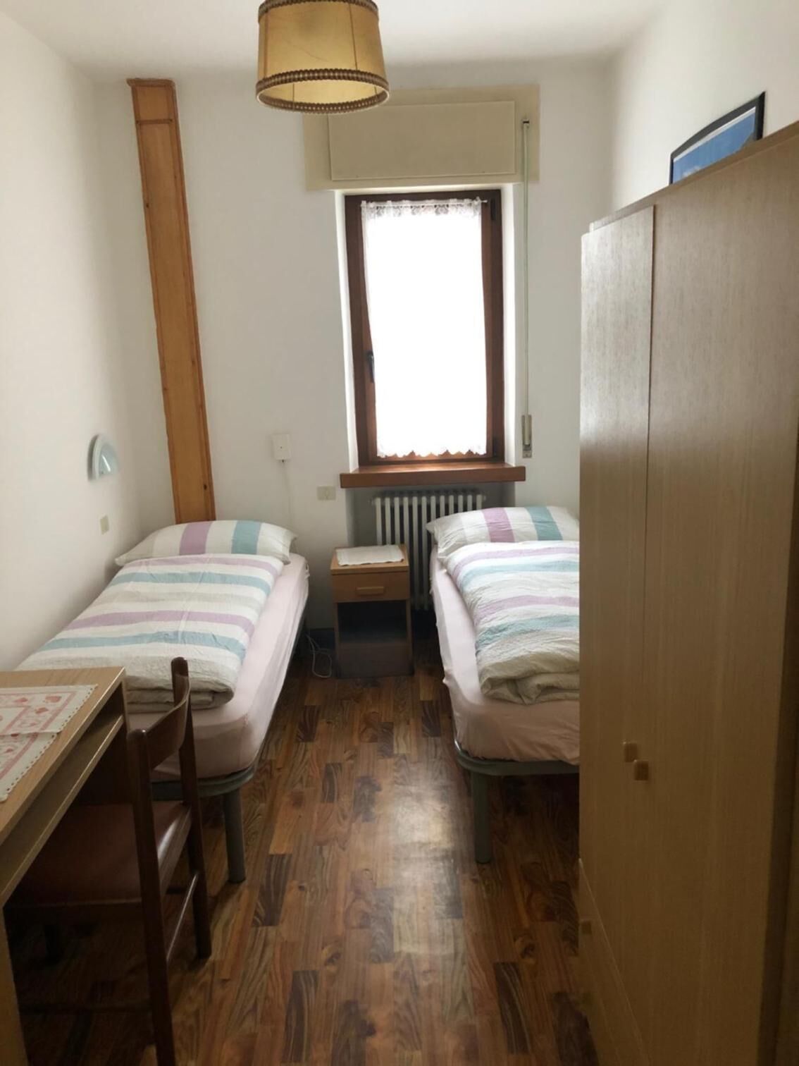 Une petite pièce, de taille modeste, comprenant deux lits simples, une armoire en bois, une table avec une chaise, un radiateur et une fenêtre avec un rideau.