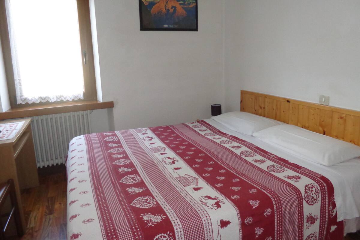 A bedroom with a wooden headboard, a patterned bedspread, a window with lace curtains, and a framed picture on the wall.