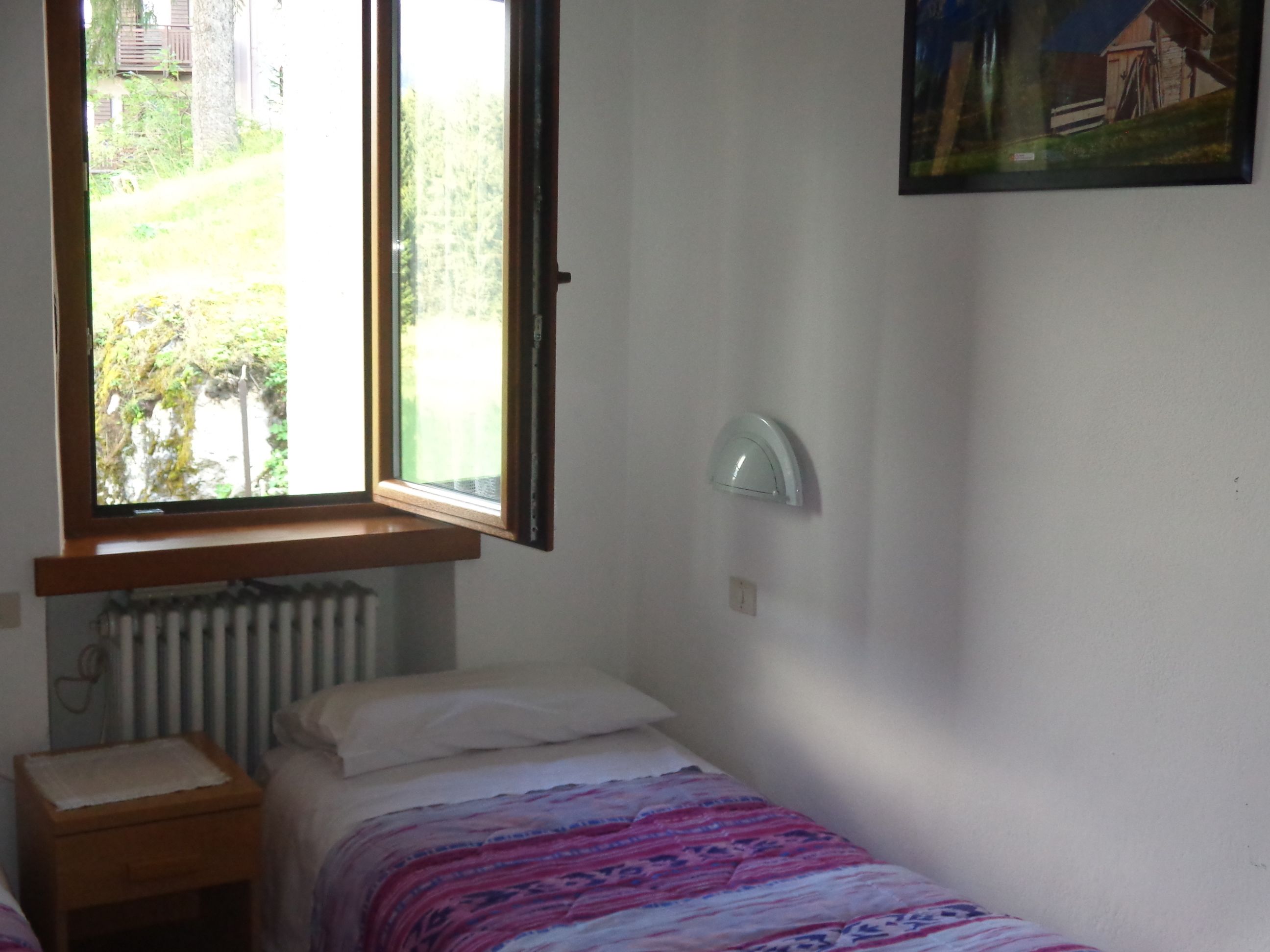 A small, simple room with a single bed, a nightstand, a window with a view of greenery, and a wall-mounted light fixture.