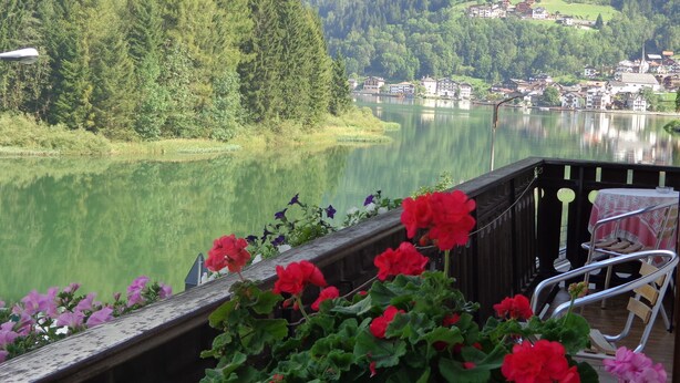 A lakeside view with a wooden deck, red flowers, and a chair. - Garnì Astor (Alleghe)