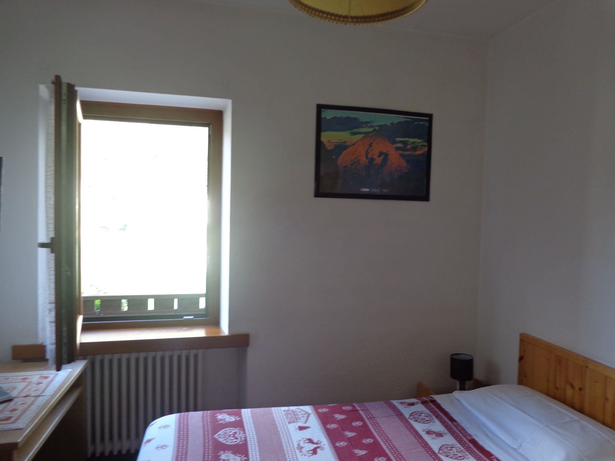 Un dormitorio con cama, ventana con cortinas, radiador, un cuadro en la pared y una mesita de noche.