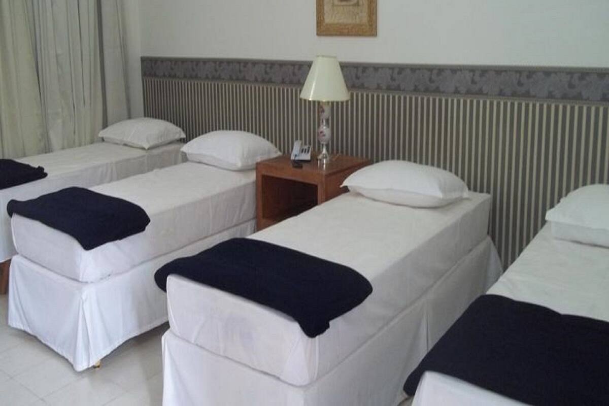 Ein Hotelzimmer mit zwei Einzelbetten, jeweils mit einer weißen Matratze und einer dunkel gefärbten Decke. Zwischen den Betten befindet sich ein Nachttisch mit einer Lampe und einem Telefon. Die Wand hinter den Betten ist mit einem Streifenmuster gestaltet.