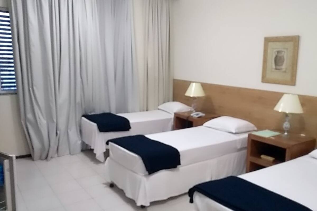 Ein Hotelzimmer mit drei Einzelbetten, jeweils mit Nachttisch und Lampe.