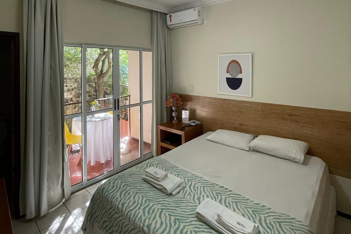 Ein Hotelzimmer mit Bett, Nachttisch, Lampe, einem gerahmten Bild und einem Fenster mit Vorhängen.