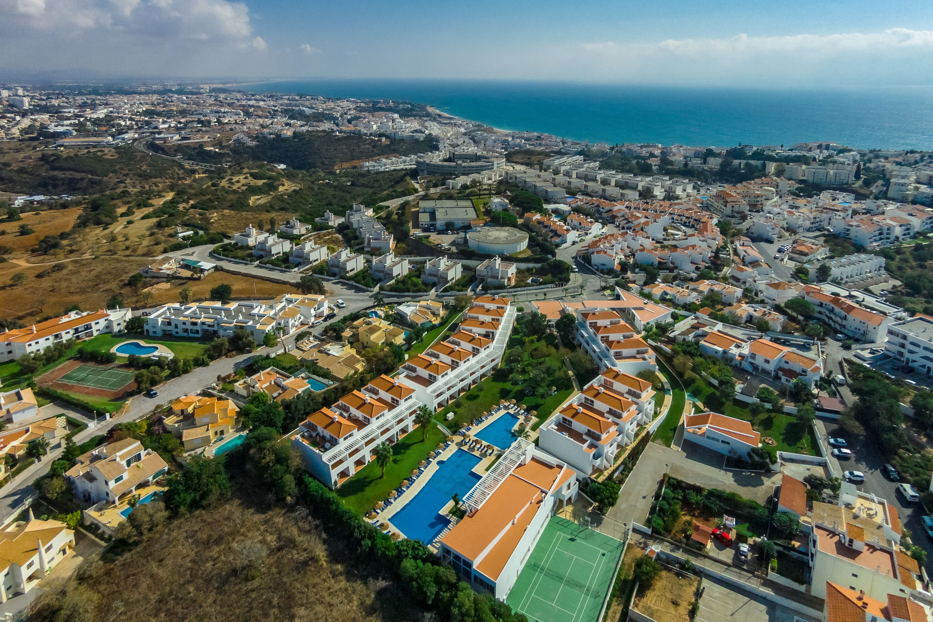 An aerial view of a coastal residential area with a large swimming pool, multiple buildings, and a clear view of the sea.