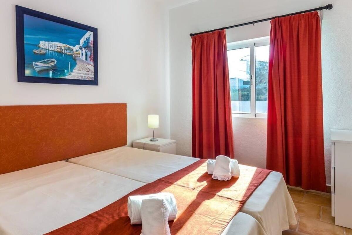 A hotel room with a bed, a nightstand, a painting of a coastal scene, and red curtains.